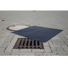 Eine schwarze Abdeckmatte liegt über einem Gully auf einem gepflasterten Untergrund.