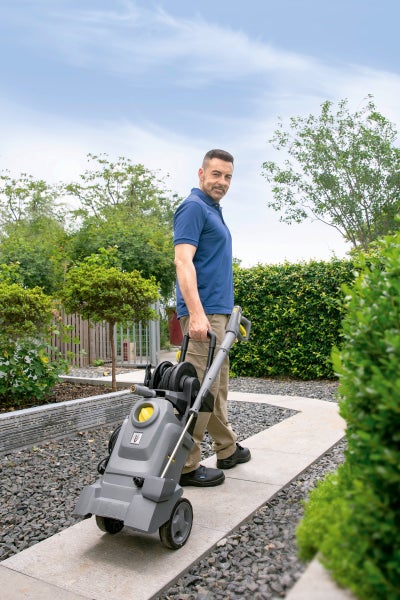 Ein Mann zieht einen fahrbaren Hochdruckreiniger mit Schlauchtrommel auf einem Gartenweg aus Steinplatten, umgeben von Kies und Hecken.