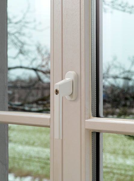 Abschließbarer weißer Fenstergriff an einem hellen Sprossenfenster mit Blick ins Grüne.