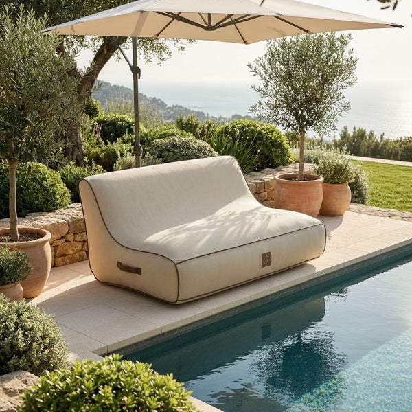 Outdoor-Lounge-Sofa für zwei Personen mit beigem Stoffbezug und brauner Paspelierung auf einer Steinterrasse am Pool unter einem Sonnenschirm.
