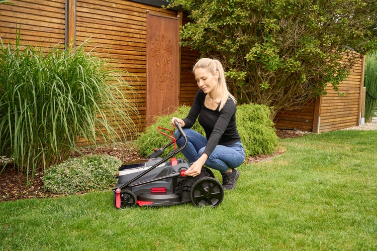 Eine Frau kniet in einem Garten neben einem Elektro-Rasenmäher auf einer grünen Rasenfläche vor einem Holzzaun und Pflanzen.