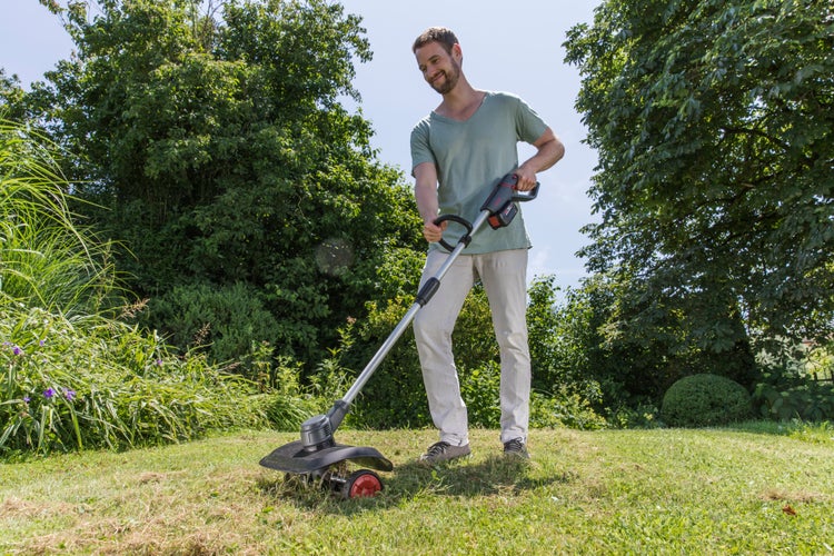 Mann trimmt Rasenkante mit einem Akku-Rasentrimmer mit Rad in einem hellen Garten.