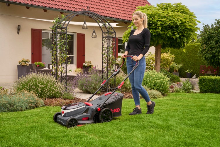 Frau mäht Rasen mit AL-KO Akku-Rasenmäher in einem hellen, gepflegten Garten mit Blumenbeeten und Wohnhaus.