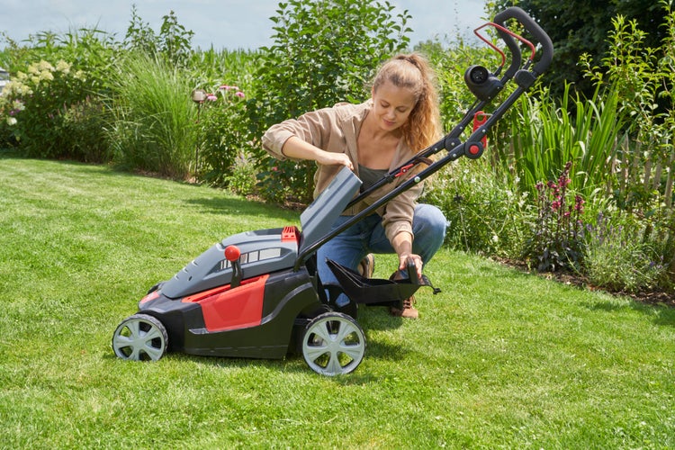 Eine Frau montiert den Grasfangkorb an einen Elektro Rasenmäher in einem sonnigen Garten.
