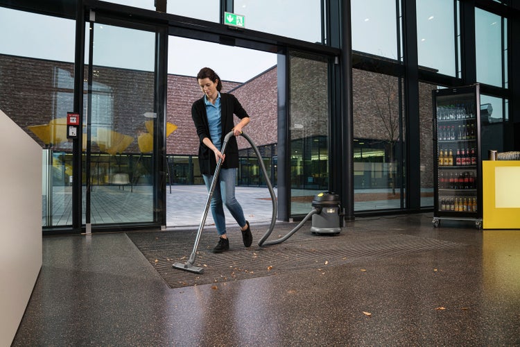 Eine Frau reinigt eine Fußmatte im modernen Eingangsbereich eines Gebäudes mit einem Kärcher Nass- und Trockensauger.