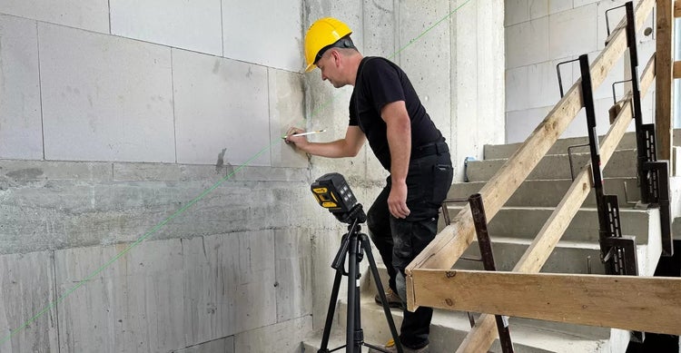 Ein Bauarbeiter mit Schutzhelm arbeitet mit einem Linienlaser an einer Wand auf einer Baustelle.