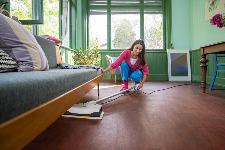 Frau reinigt mit einem Kärcher Dampfreiniger unter einem grauen Sofa in einem hellen Zimmer mit grünen Wänden und Holzboden.