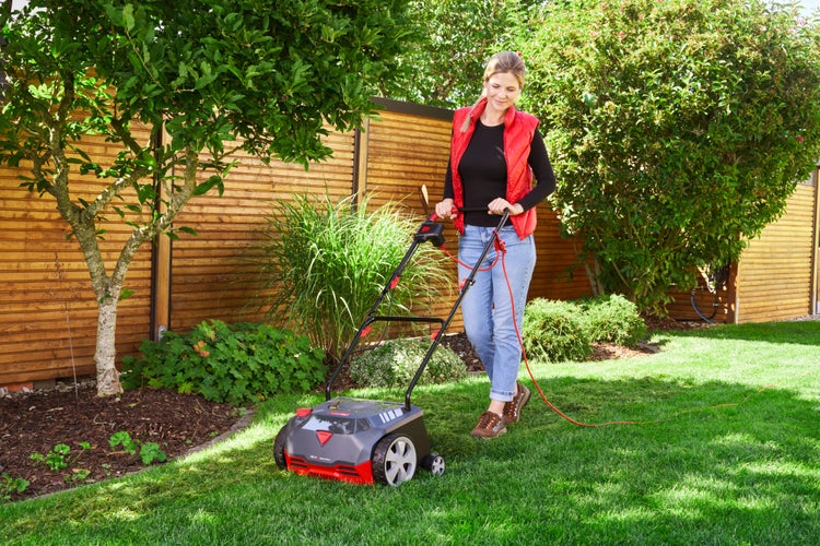 Eine Frau bedient einen Elektro-Vertikutierer auf einer grünen Rasenfläche im Garten vor einem Holzzaun.