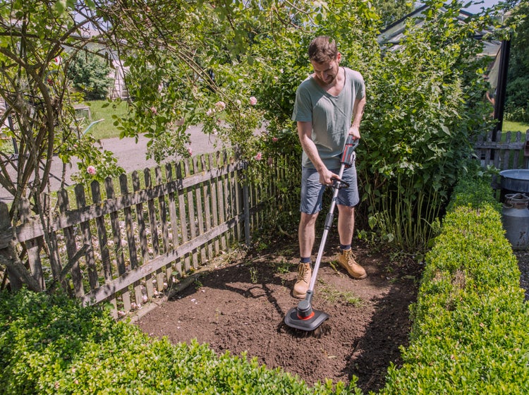 Ein Mann benutzt eine motorisierte Gartenfräse in einem hellen Garten, um Erde neben einem Holzzaun und einer Hecke umzugraben.