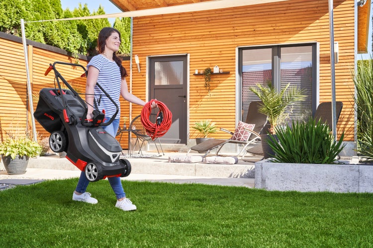 Eine Frau trägt einen elektrischen Rasenmäher der Marke AL-KO an einer Holzfassade vorbei im Garten.