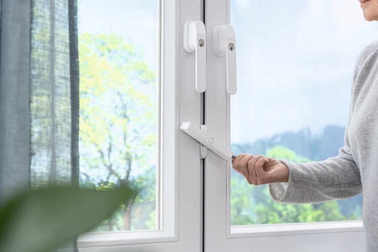 Eine Person betätigt eine weiße Fenstersicherung an einem weißen Fensterrahmen mit abschließbaren Fenstergriffen.
