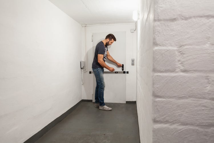 Ein Mann bedient einen schwarzen Panzerriegel an einer weißen Tür in einem hellen Kellerraum mit weißen Wänden und grauem Boden.