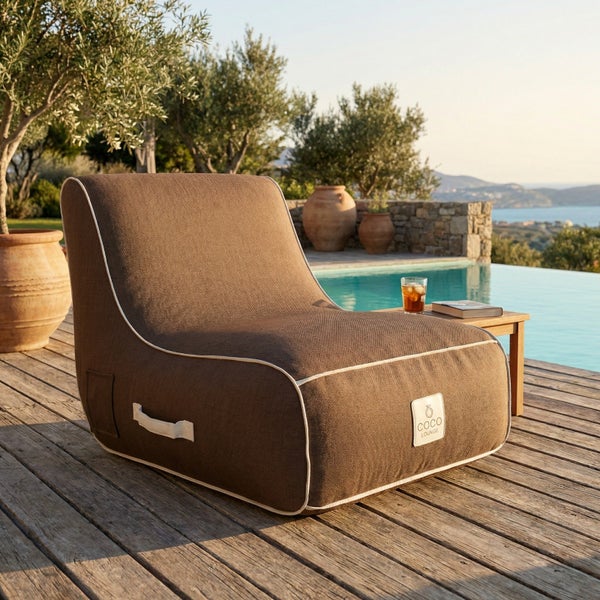 Moderner Lounge-Sesselsitzsack in Braun mit weißem Rand und Tragegriff auf einer sonnigen Holzterrasse am Pool. COCO LOUNGE Logo.
