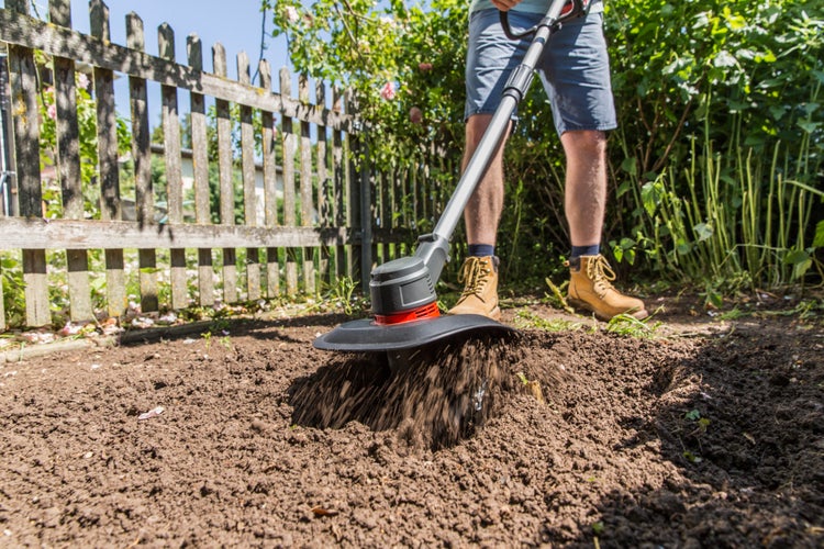 Person benutzt eine Gartenfräse zum Umgraben von Erde in einem Garten mit Holzzaun.
