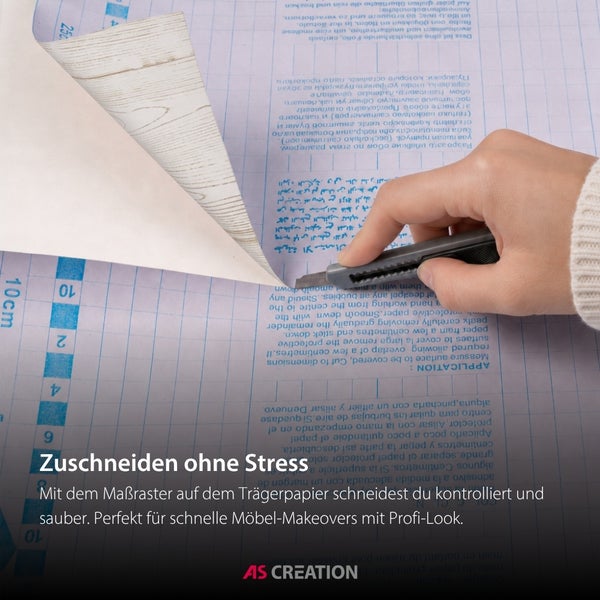 Zuschneiden von selbstklebender Möbelfolie in Holzoptik mit einem Cuttermesser entlang des Rasters auf dem Trägerpapier. AS CREATION Logo.