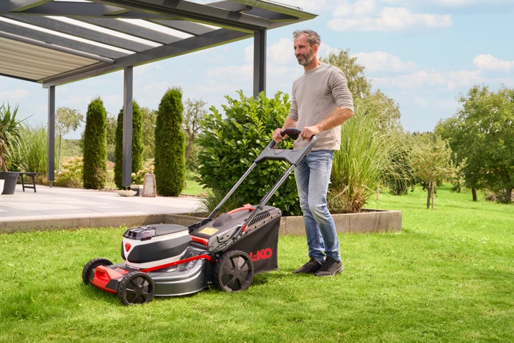 Mann mäht den Rasen mit einem AL-KO Akku-Rasenmäher in einem gepflegten Garten neben einer Terrasse mit Überdachung.