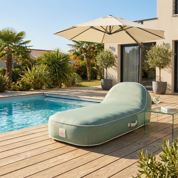 Eine mintgrüne Outdoor-Liege in Sitzsack-Optik auf einer Holzterrasse neben einem Pool, geschützt durch einen großen Sonnenschirm.
