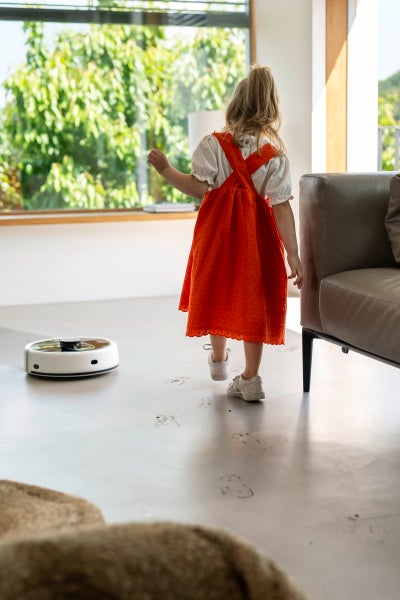 Ein Kind in einem roten Kleid steht in einem hellen Raum, während ein Staubsaugerroboter den Boden reinigt.