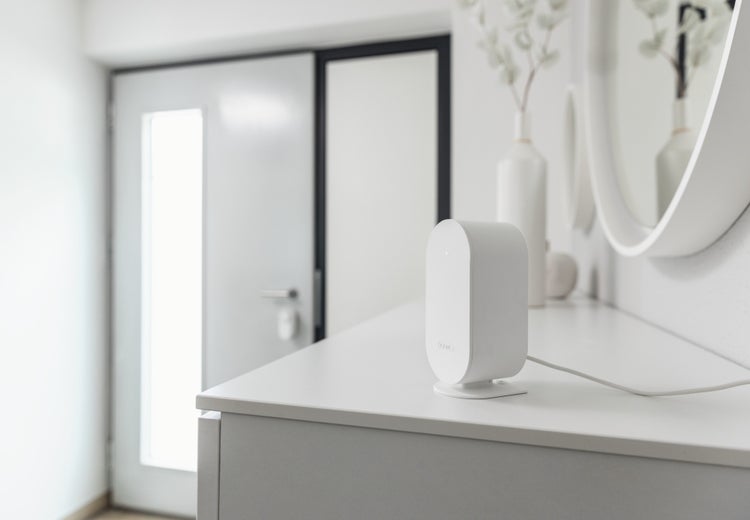 Ein weißes ABUS Smart-Home-Gerät steht auf einem weißen Sideboard in einem hellen, modernen Flur mit weißer Tür und Spiegel im Hintergrund.