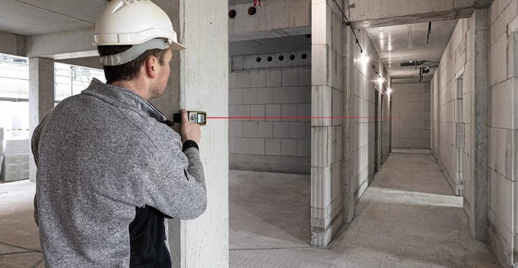 Ein Bauarbeiter mit Schutzhelm benutzt einen Laser-Entfernungsmesser auf einer Baustelle, um eine rote Laserlinie an einer Wand auszurichten.