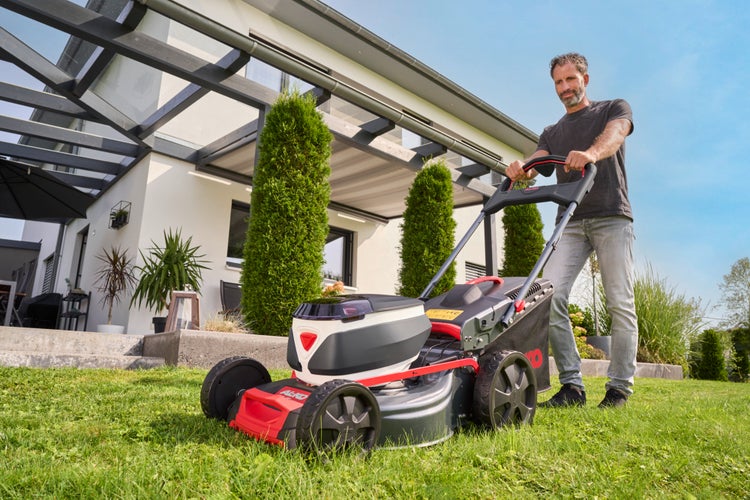 Ein Mann mäht den Rasen in einem modernen Garten mit einem AL-KO Akku-Rasenmäher vor einem hellen Wohnhaus.