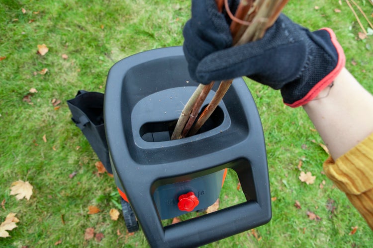 Eine Hand mit Gartenhandschuh führt dünne Äste in die Einfüllöffnung eines elektrischen Gartenhäckslers ein.