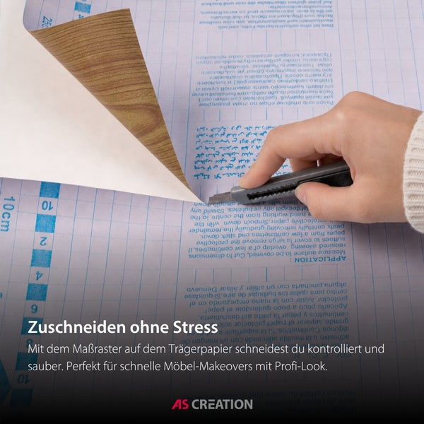 Eine Hand schneidet eine selbstklebende Möbelfolie in Holzoptik mit einem Cuttermesser entlang des Rasters auf der Rückseite zu. AS Creation Logo.