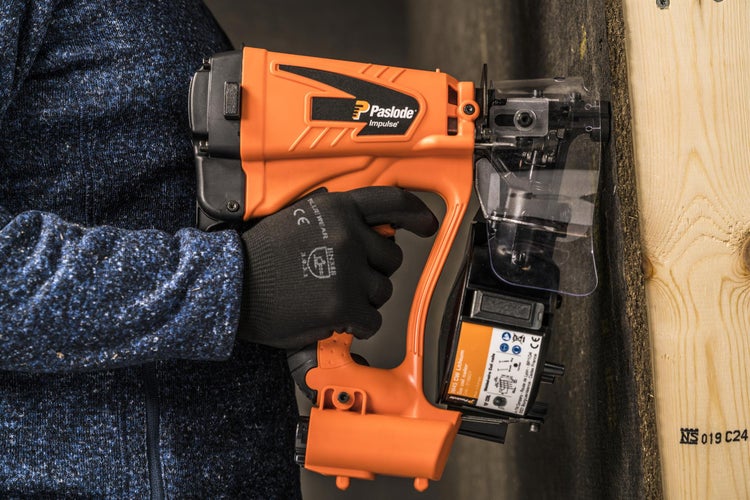 Eine Person bedient einen orangefarbenen Paslode Impuls Coilnagler an einer Holzwand.
