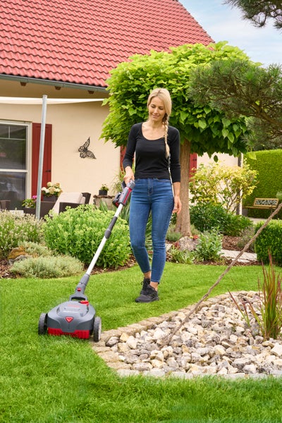 Frau mäht Rasen mit kompaktem Akku-Rasenmäher in einem gepflegten Garten mit Blumenbeeten und Haus.
