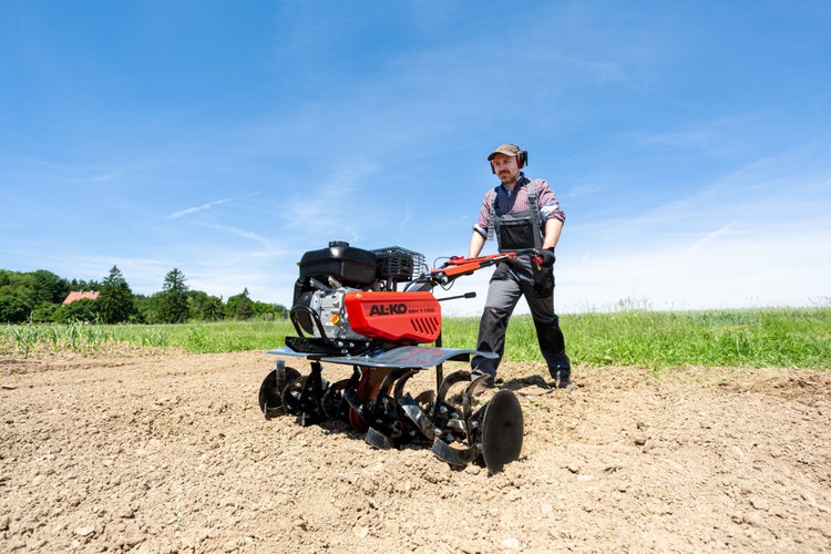 Ein Mann verwendet eine AL-KO Benzin-Motorhacke MH 1150 zur Bodenbearbeitung auf einem Feld unter blauem Himmel.