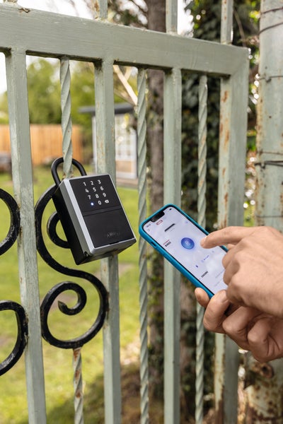 Elektronischer Schlüsseltresor von ABUS an einem Metalltor. Eine Hand steuert den Tresor über eine App auf einem Smartphone.