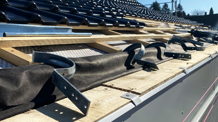 Dachkonstruktion mit Dachziegeln, Holzlatten, Unterspannbahn und vormontierten Rinnenhaltern aus Metall auf einer hellen Baustelle.