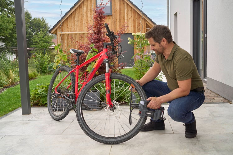 Ein Mann pumpt mit einem batteriebetriebenen Kompressor den Vorderreifen eines roten Mountainbikes auf einer hellen Terrasse im gepflegten Garten auf