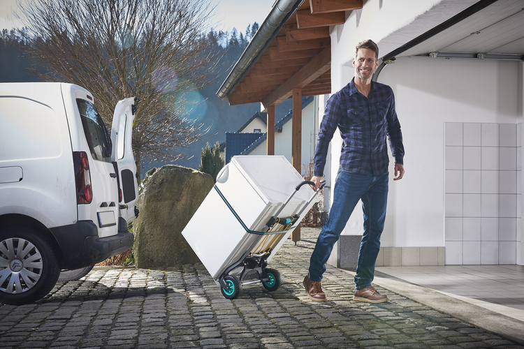 Mann nutzt eine klappbare Sackkarre zum Transport einer großen weißen Box von einem Transporter in eine Garage auf gepflastertem Untergrund.