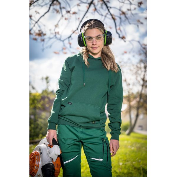 Frau trägt grüne Arbeitskleidung mit Schutzbrille und Kopfhörern im Freien.