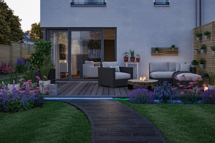 Moderne Terrasse mit Polyrattan-Gartenmöbeln, Holzdielen, gepflastertem Weg, LED-Außenbeleuchtung und verschiedenen Pflanzen bei Dämmerung.