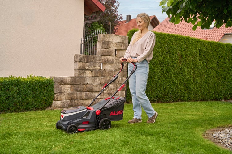 Eine Frau kniet in einem hellen Garten und stellt die Schnitthöhe eines AL-KO Rasenmähers auf einem Rasen ein.