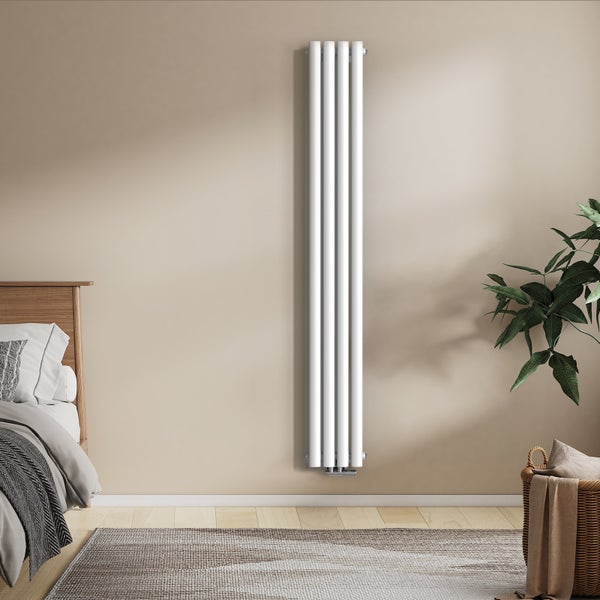 Vertikaler Designheizkörper mit vier Rundrohren an einer Wand im Schlafzimmer mit Bett und Zimmerpflanze.