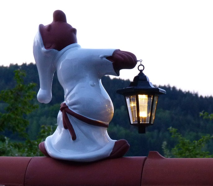 Gartenfigur Wichtel im Nachtgewand mit leuchtender Laterne auf einem Dachziegel vor Waldhintergrund.