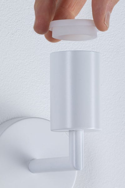 Eine Hand setzt einen weißen lichtdurchlässigen Ring auf eine weiße zylindrische Lampenfassung einer Wandleuchte.