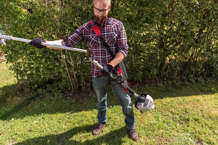 Ein Gärtner montiert einen AL-KO Motorsensen Aufsatz an einen Schaft vor einer Hecke.