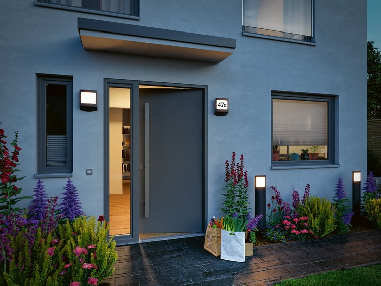 Moderner Hauseingang mit anthrazitfarbener Haustür, Wandleuchte mit Hausnummer, Wegeleuchten im Blumenbeet und Einkaufstüten vor der Tür.
