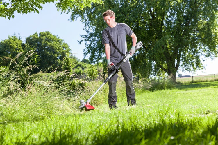 Ein Mann mit Schutzausrüstung verwendet einen Akku-Rasentrimmer, um hohes Gras auf einer Wiese an einem sonnigen Tag zu schneiden.