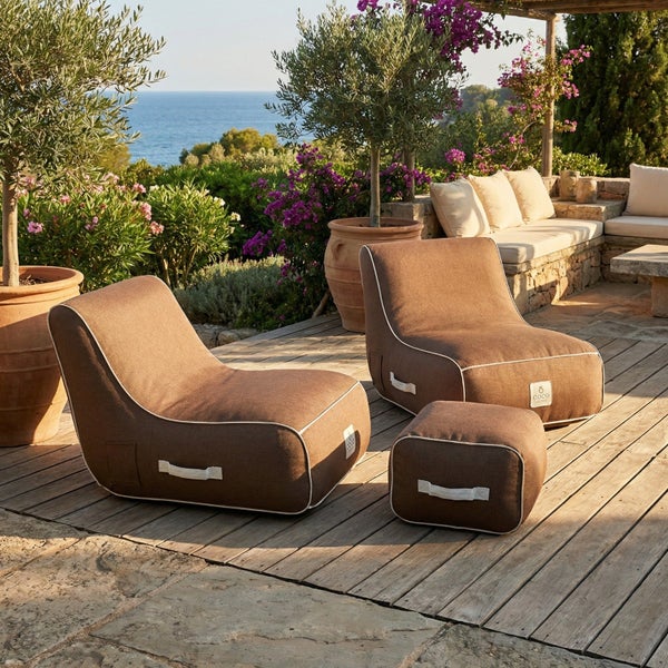 Zwei braune Outdoor-Sitzsäcke und ein Hocker aus Stoff auf einer hellen Holzterrasse mit Meerblick und mediterraner Bepflanzung.