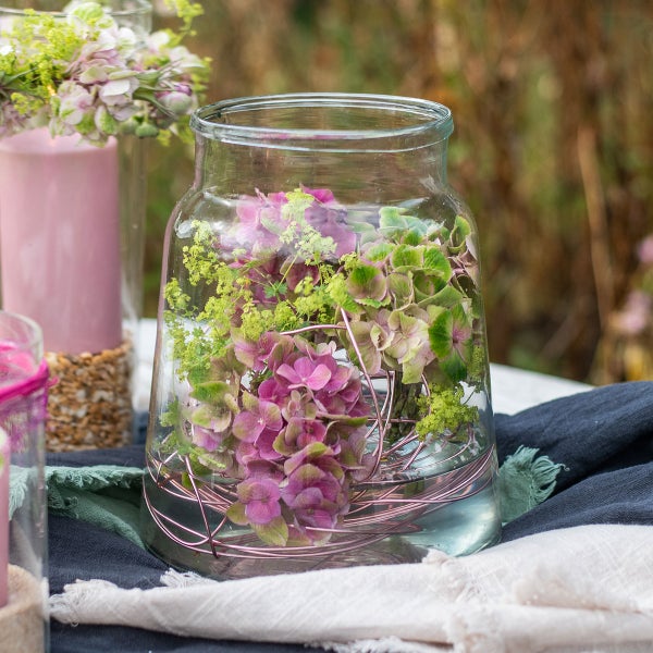 Transparentes Glasgefäß als Vase mit rosa und grünen Hortensien sowie rosa Aluminiumdraht auf einem Tisch im Freien.
