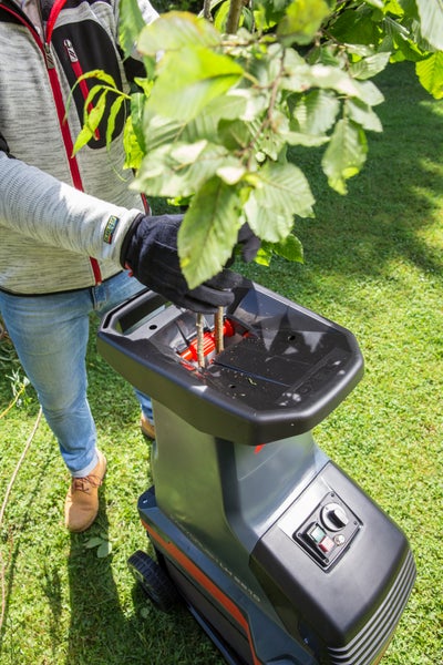 Eine Person führt mit Handschuhen beblätterte Äste in die Einfüllöffnung eines elektrischen Gartenhäckslers auf einer Rasenfläche ein.