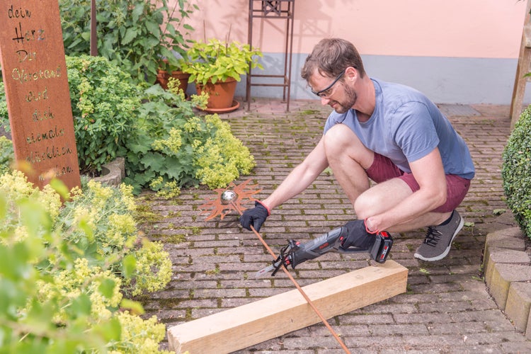 Ein Mann schneidet mit einer Akku-Säbelsäge einen Metallstab auf einem Holzbalken im Garten.