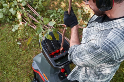 Eine Person führt mit Handschuhen und Gehörschutz einen Ast in einen Gartenhäcksler ein.