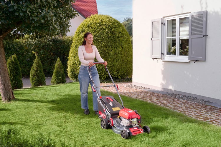 Eine Frau mäht den Rasen mit einem AL-KO Benzin-Rasenmäher in einem hellen, gepflegten Garten neben einem Haus mit grauen Fensterläden.