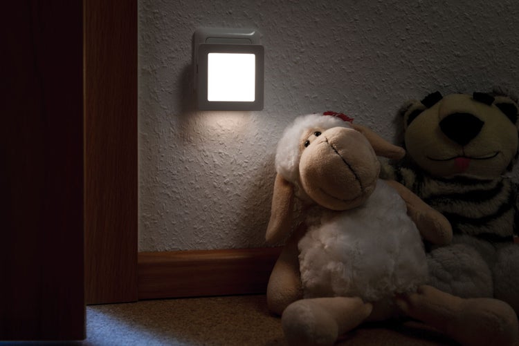 Quadratisches LED-Nachtlicht für die Steckdose, leuchtend an der Wand neben Plüschtieren in einem abgedunkelten Kinderzimmer.